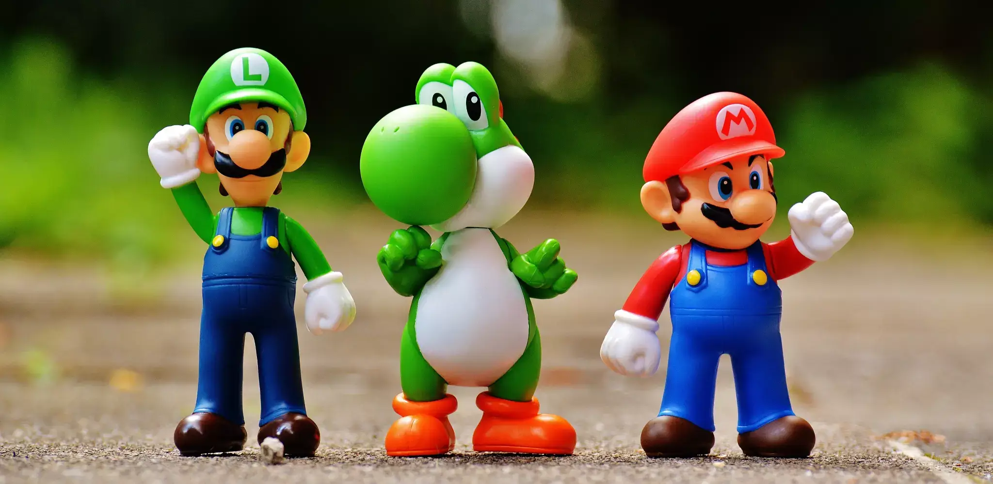 Luigi, Yoshi, and Mario standing side by side on a cement surface, with a blurred backdrop of what appears to be a park.